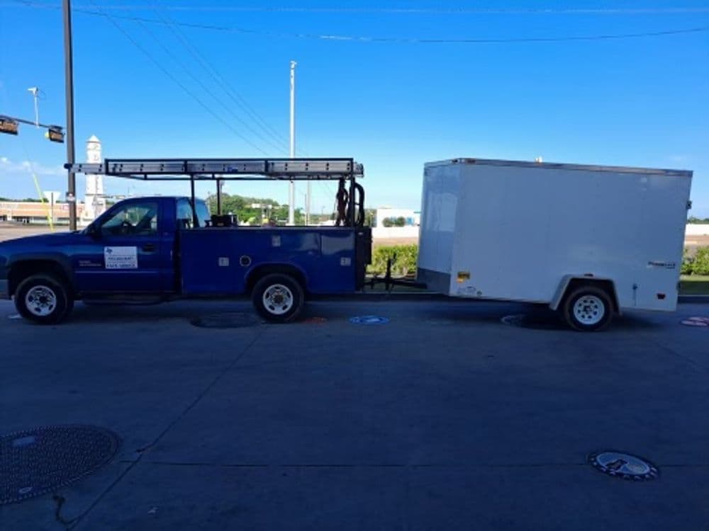 Blue utility truck with ladder rack towing a white enclosed trailer on sunny day.