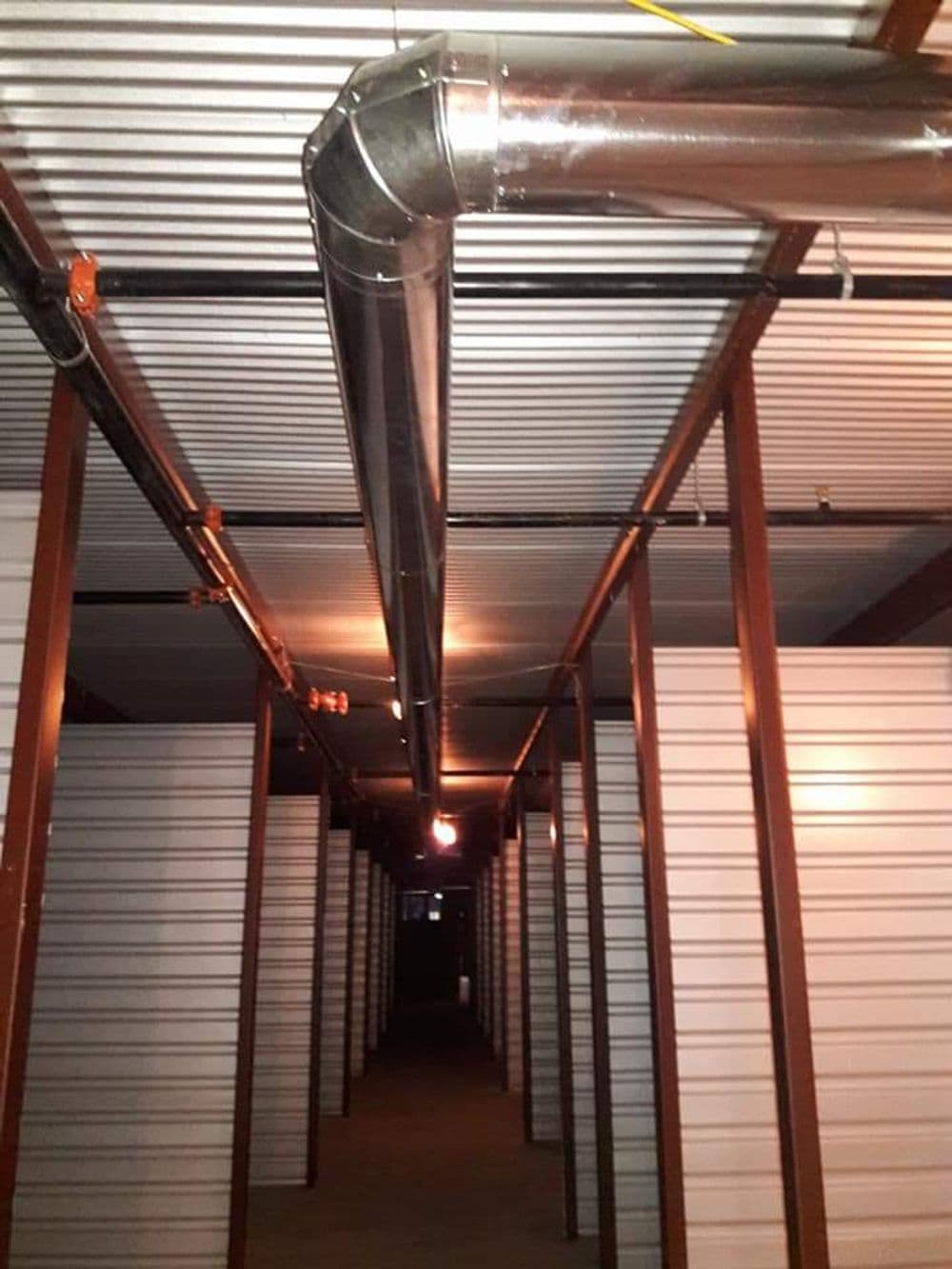 Storage facility interior featuring metal piping and illuminated corridor between units.