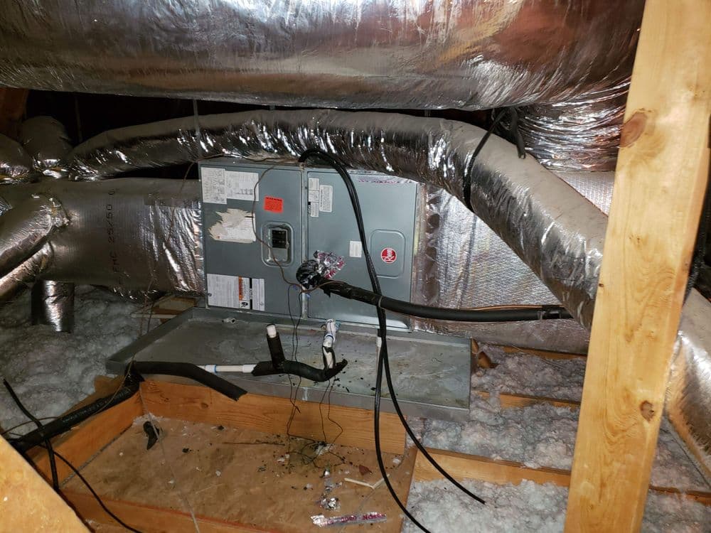 HVAC unit in attic with ductwork and thermal insulation visible on wooden beams.