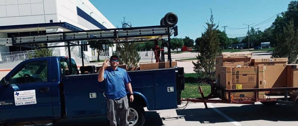 Man gesturing by a blue pickup truck with boxes on a trailer, in a parking lot.