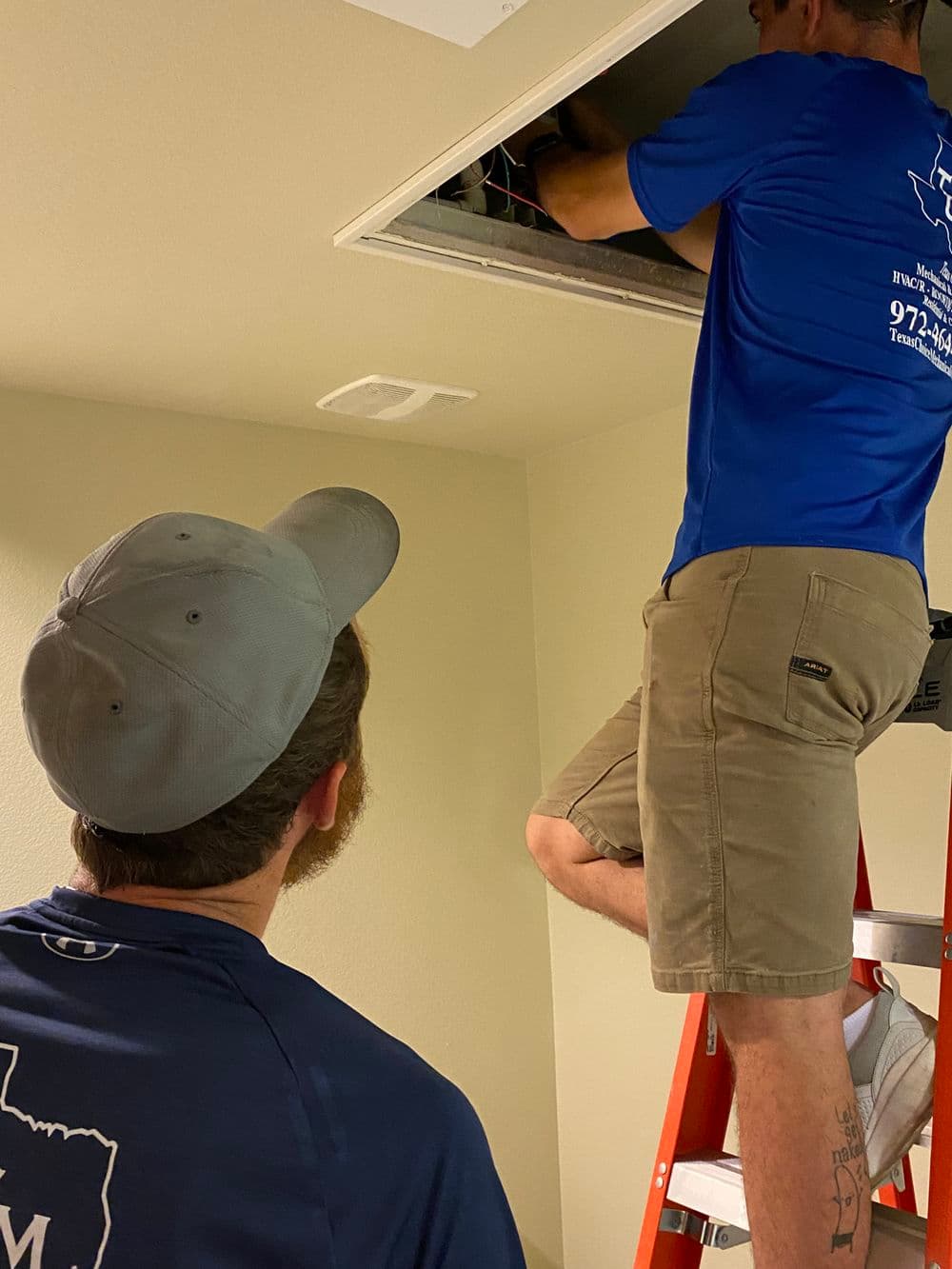 Technician working on ceiling while another observes from below on a ladder.
