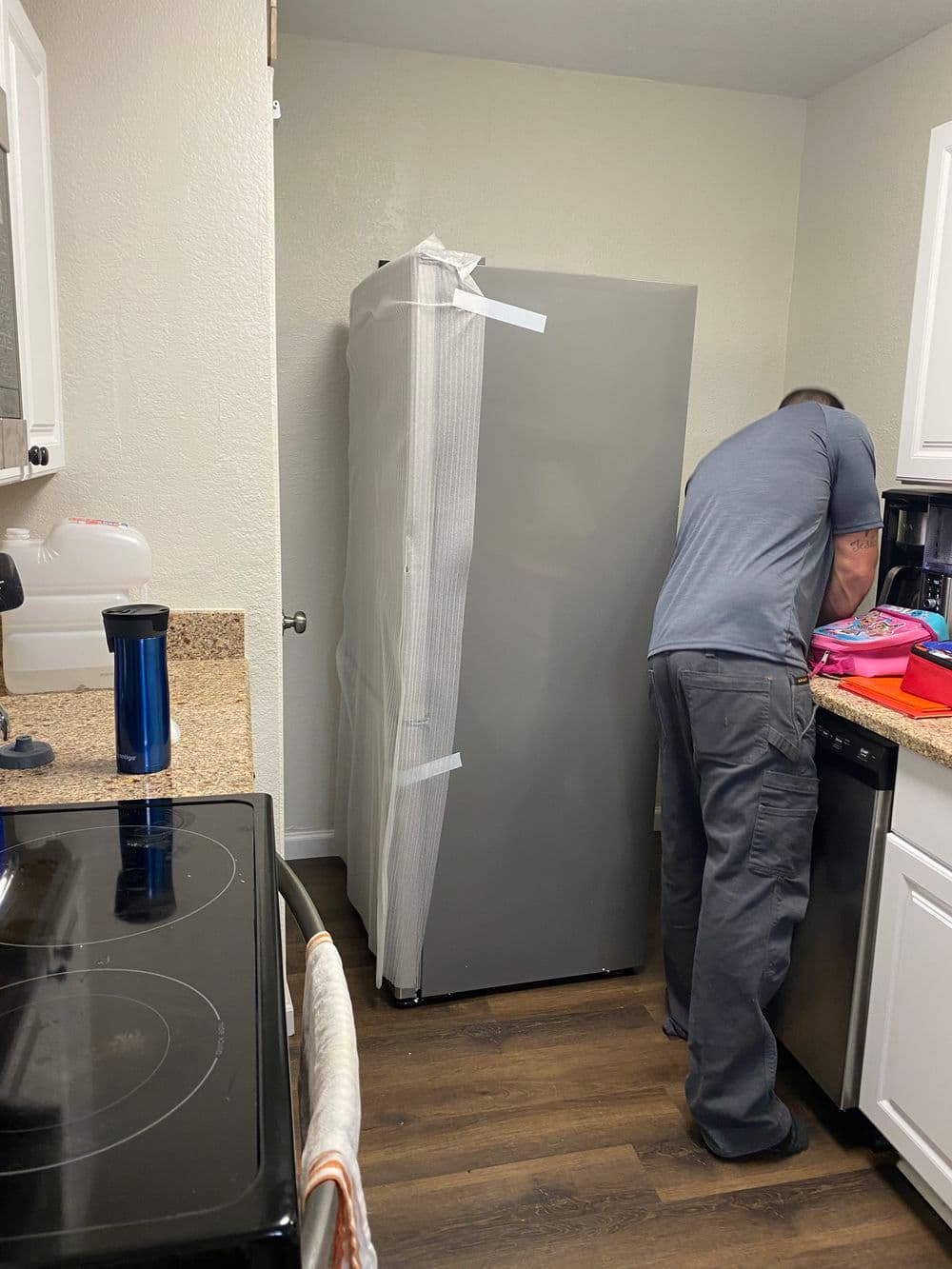 Man unpacking a new refrigerator in a modern kitchen with white cabinets and dark flooring.