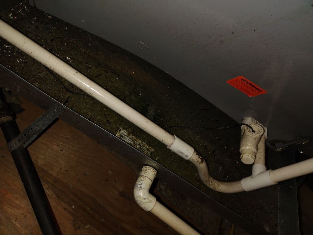 Close-up of plumbing pipes with warning label under a sink, showing dirt and potential leaks.