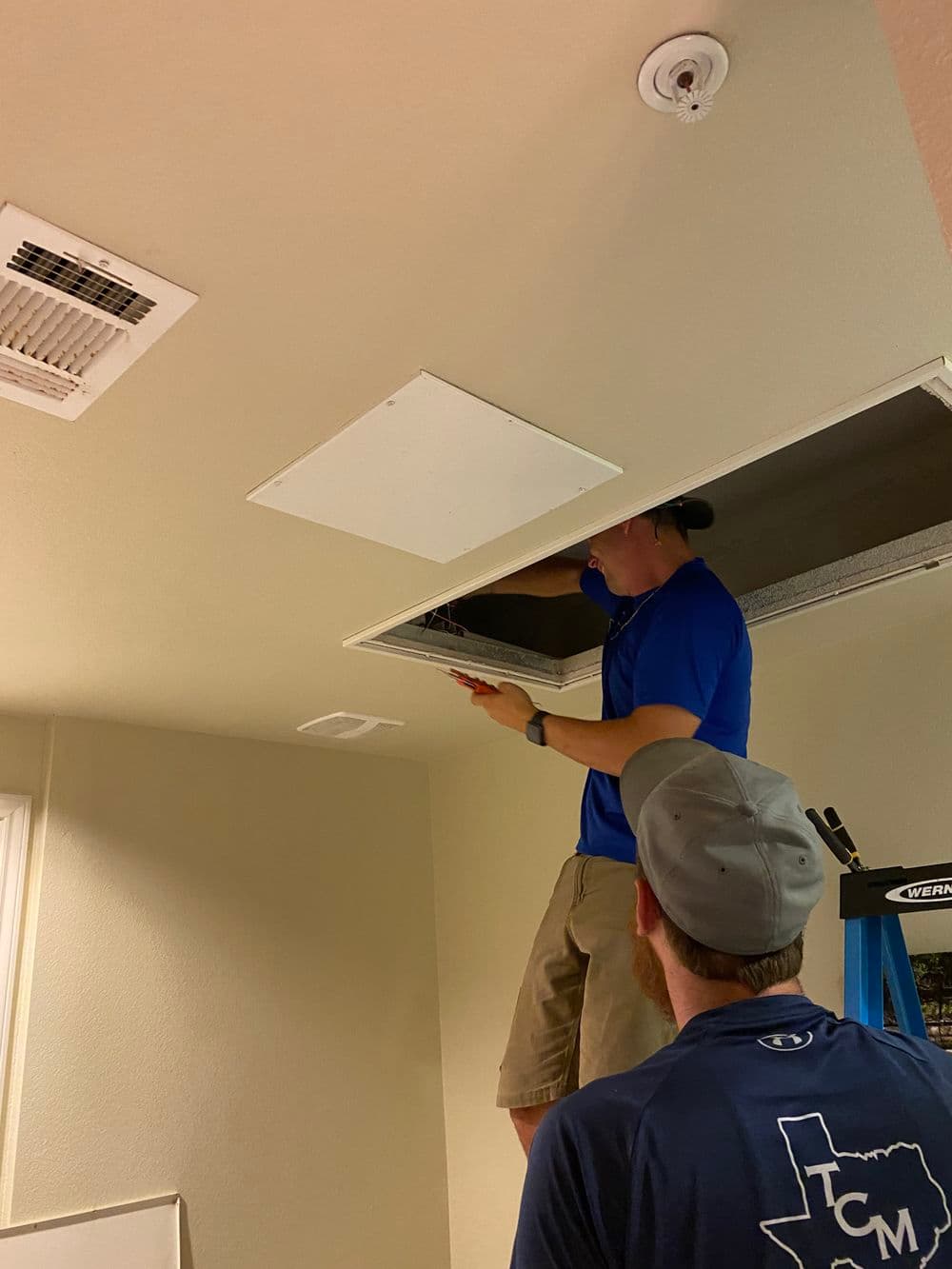 Man working on ceiling access panel with tools, another person observing from below.