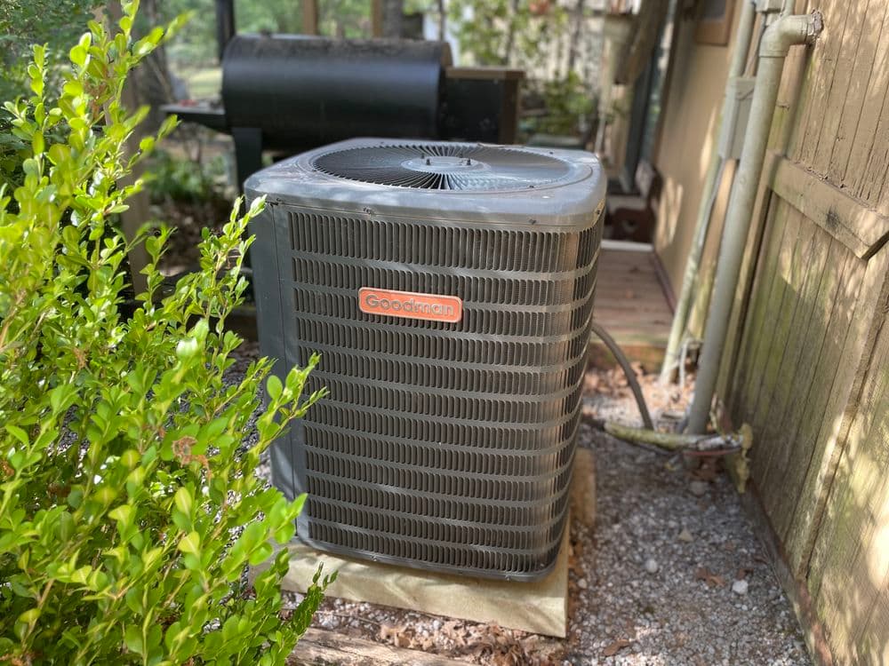 Goodman air conditioning unit beside a house, surrounded by greenery and gravel.