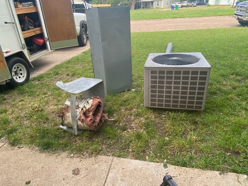 Disassembled air conditioning unit and metal scrap on grass near parked vehicles.